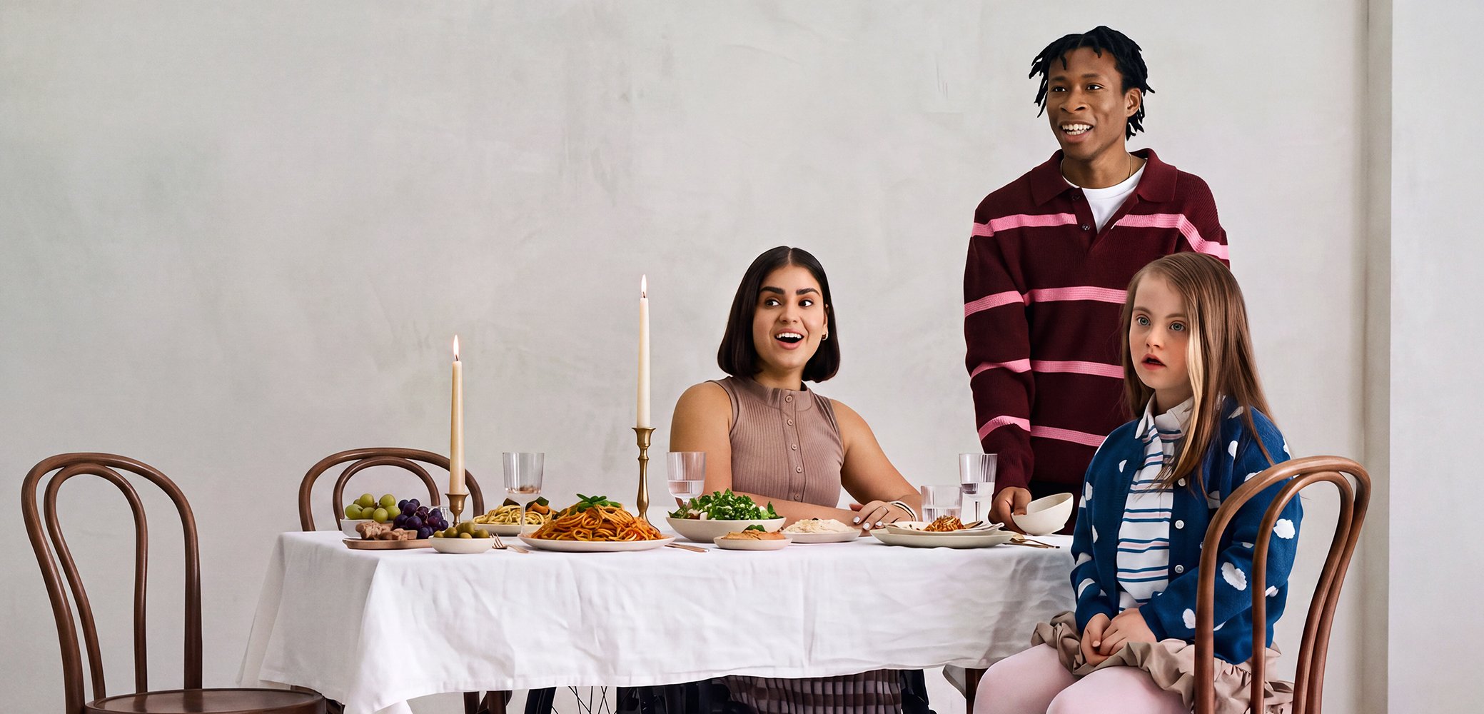 Three diverse young adults at a dining table with food, candles, and empty chairs against a plain light wall background.