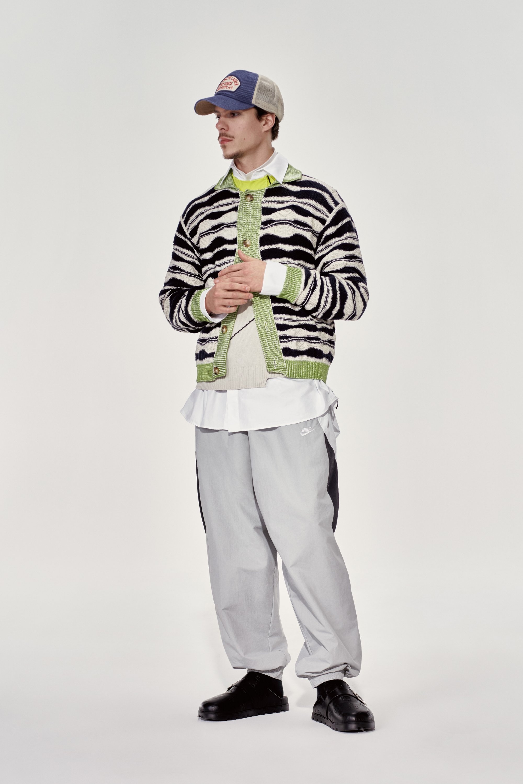 Un joven está de pie, vistiendo un cárdigan a rayas negro y blanco sobre una camiseta verde y una blusa blanca en capas, combinado con pantalones grises y zapatos negros.