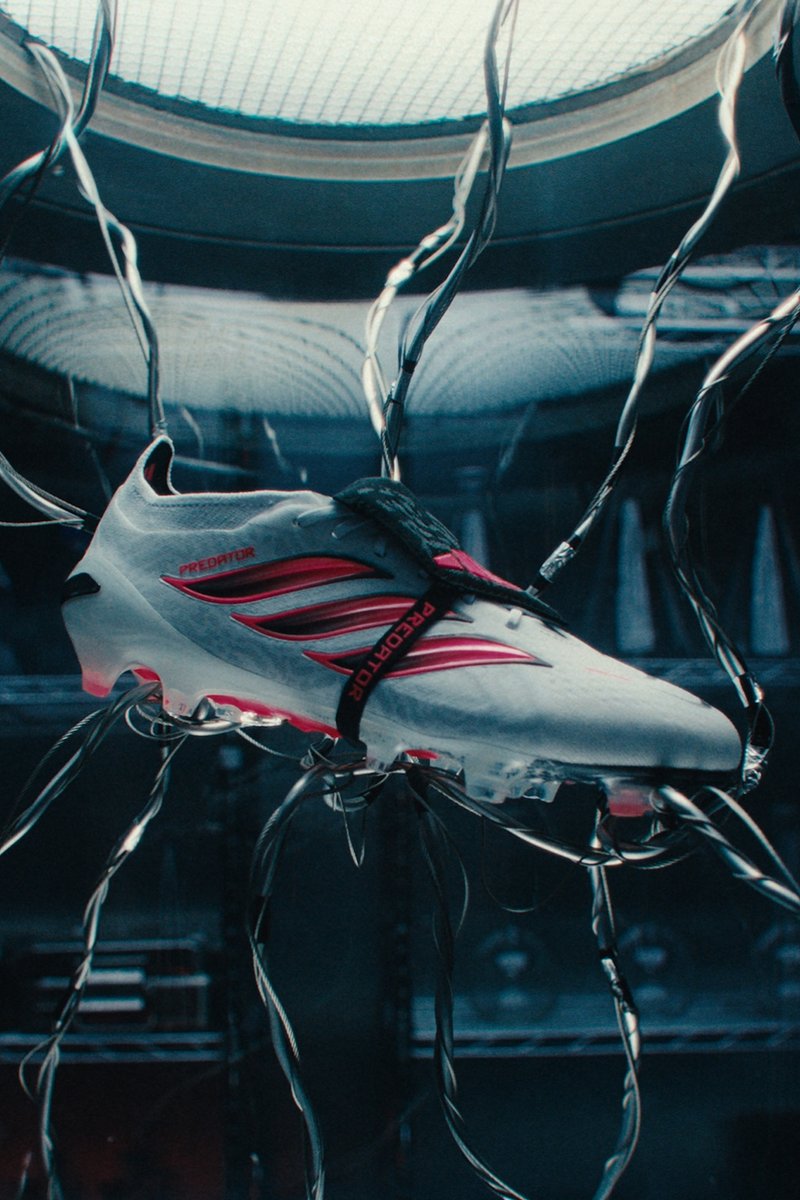 Weißer und roter Adidas Predator Fußballschuh, an Kabeln aufgehängt in einem dunklen futuristischen Laborumfeld.
