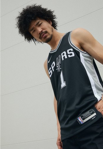 Jeune homme aux cheveux bouclés portant un maillot noir des Spurs, regardant directement l'appareil photo devant un fond gris clair texturé.