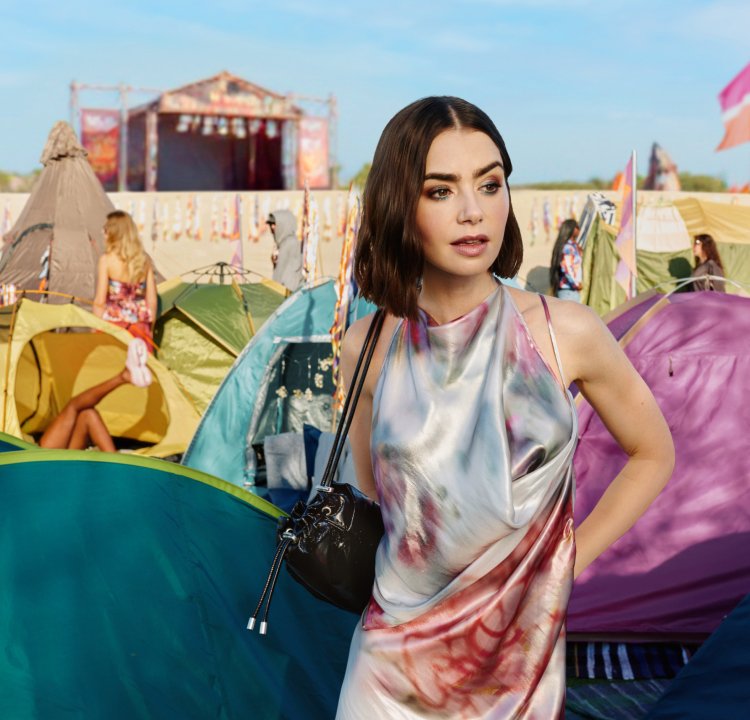 Jeune femme avec un petit sac noir se tient parmi des tentes colorées lors d'un festival en plein air avec une scène et des drapeaux en arrière-plan.