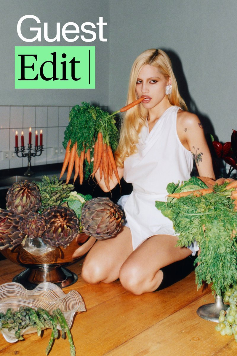 Una mujer con un vestido blanco se arrodilla sobre una mesa de madera, sosteniendo grandes racimos de zanahorias y verduras, rodeada de productos coloridos.