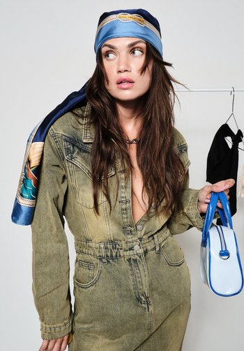 Frau mit langen braunen Haaren, die eine verblasste olivgrüne Denimjacke und ein blau gemustertes Kopftuch trägt, hält eine weiße und blaue Handtasche und schaut seitlich.