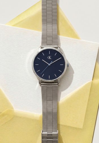 Montre-bracelet à bracelet en maille argentée avec un cadran noir minimaliste affichant 10:09:38, reposant sur des feuilles de papier translucides blanches et jaunes.