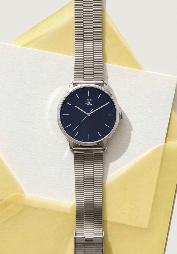 Silver mesh strap wristwatch with a black minimalist dial displaying 10:09:38, placed on white and yellow translucent paper sheets.