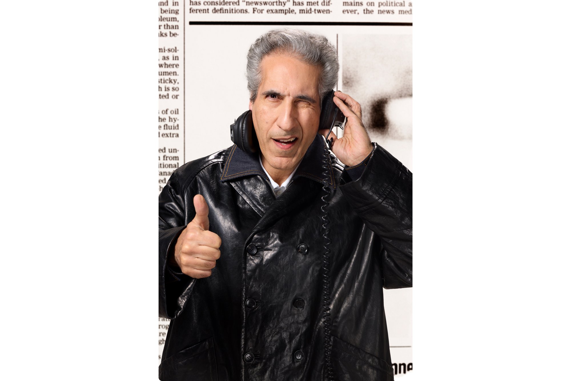 Un uomo sorridente in un cappotto di pelle nera fa il pollice in alto indossando cuffie e tenendo un telefono, con uno sfondo di giornali.