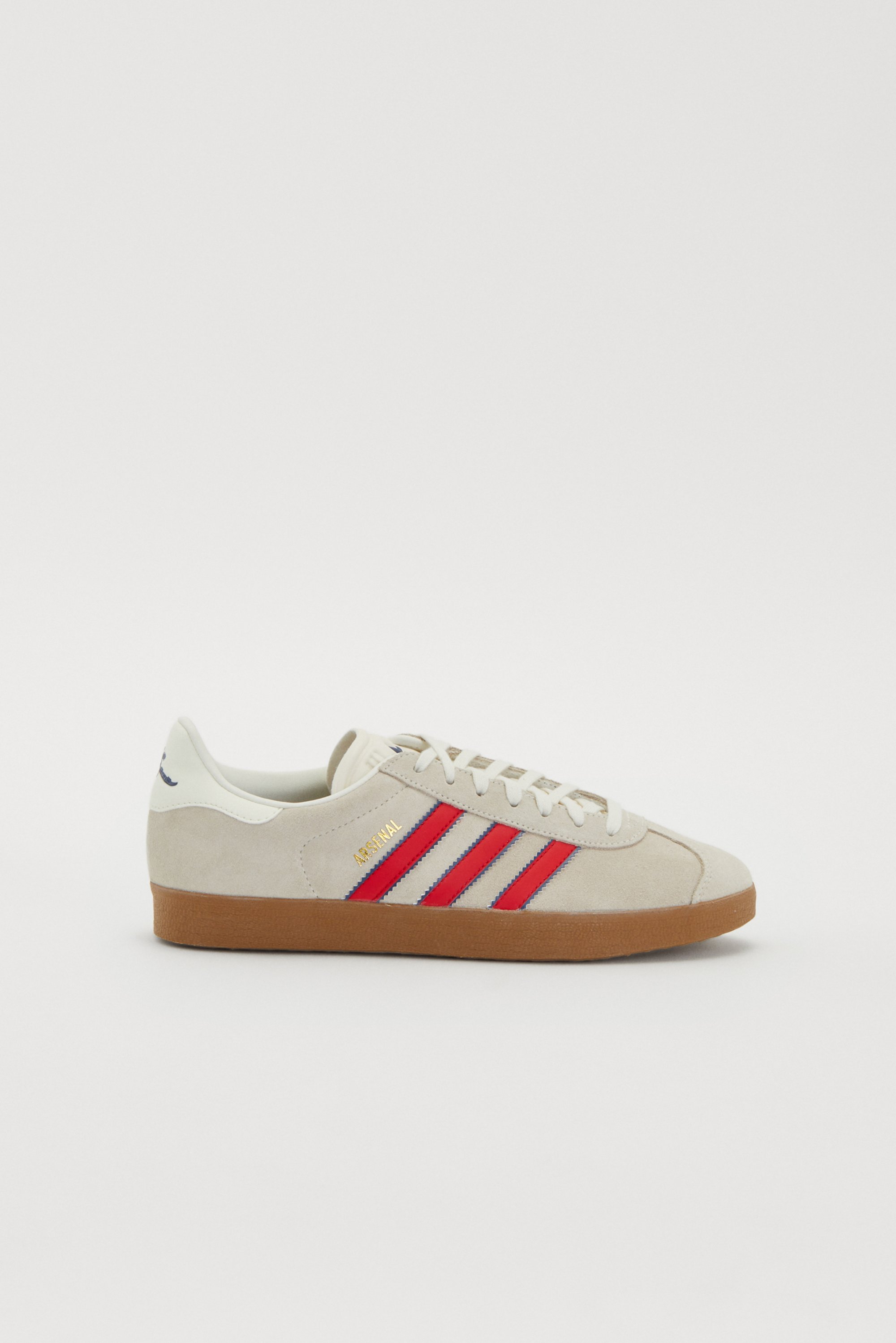 Grijze sneaker met rode en marineblauwe strepen, met een beige suède bovenwerk en een bruine rubberen zool. "Arsenal" logo zichtbaar aan de zijkant.