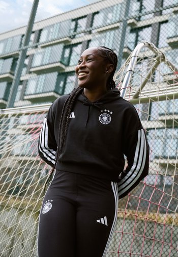 Smaidīga sieviete melnā sporta tērpā ar baltām svītrām stāv atslābināta pie futbola vārtu konstrukcijas pilsētas laukumā ar ķēžu žogu.