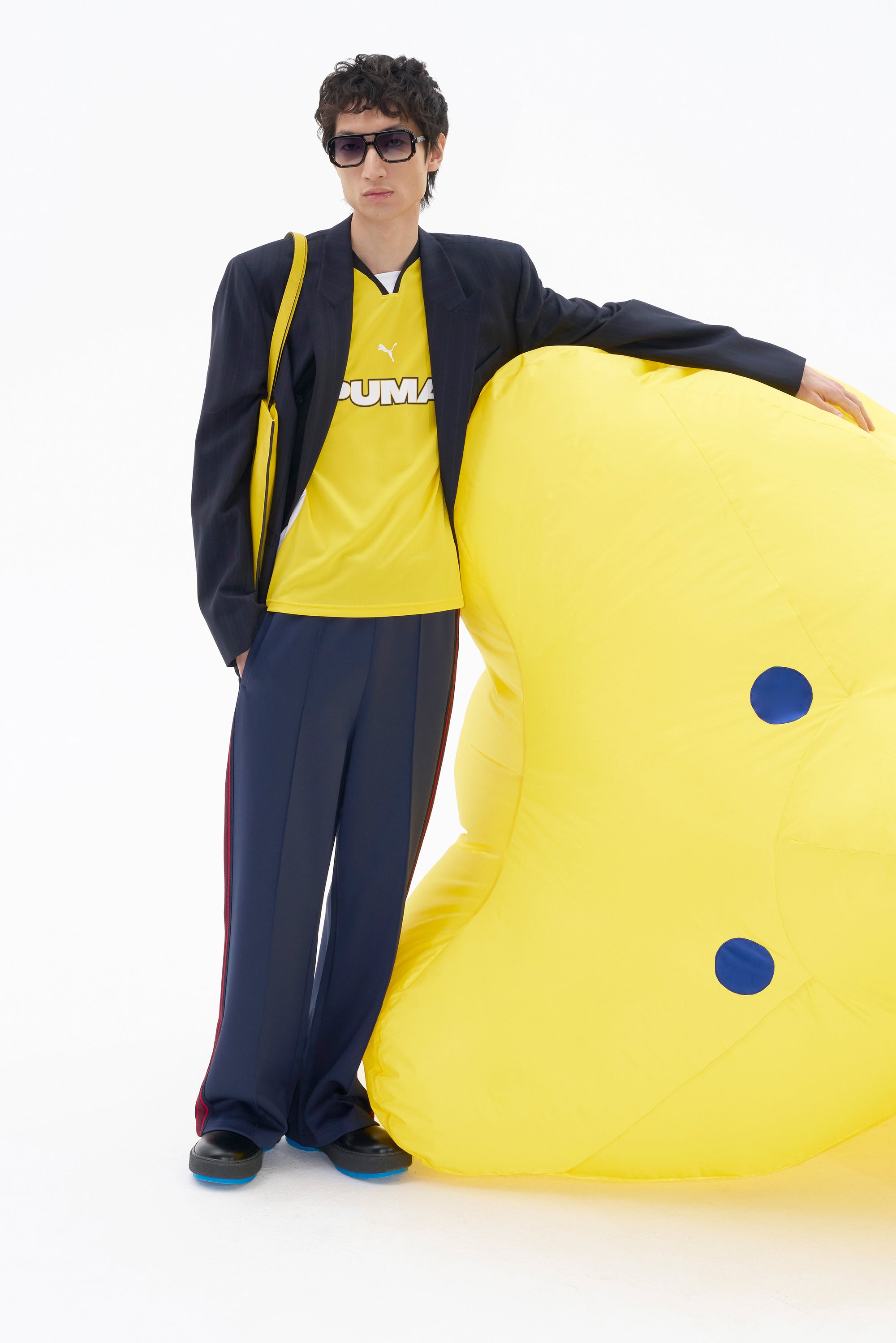 Una persona con una camiseta amarilla de Puma y un traje oscuro posa con un gran objeto inflable amarillo. El fondo es blanco, creando un aspecto minimalista.