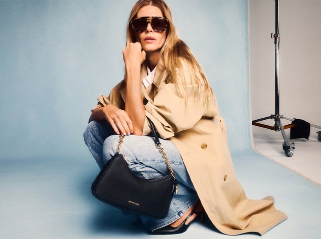 Femme accroupie portant un trench-coat beige, un jean bleu, des talons noirs, des lunettes de soleil oversize, tenant un sac à main noir Michael Kors dans un studio photo.