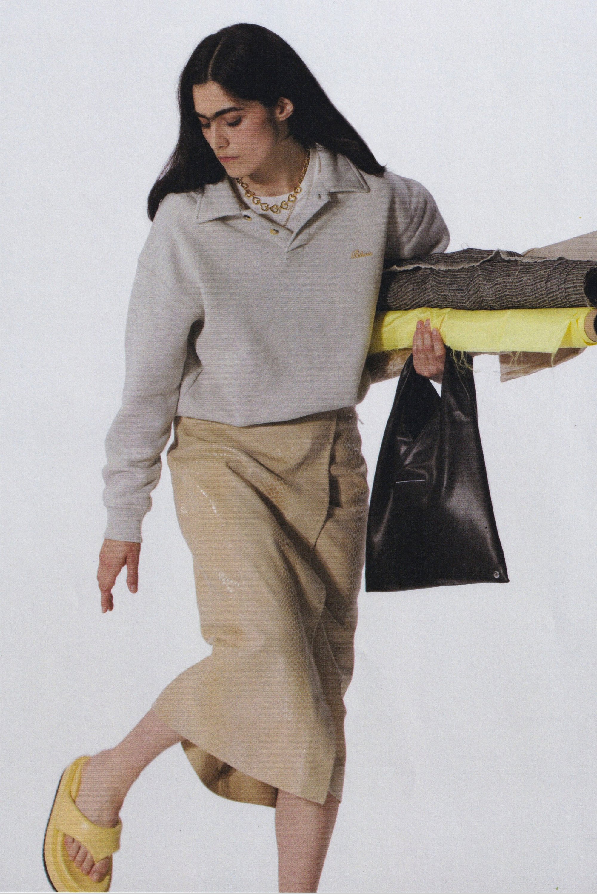 Lichtgrijze sweatshirt met kraag, beige glanzende leren rok, zwarte tas, gele opgerolde stof. Gele sandalen met dikke zool.