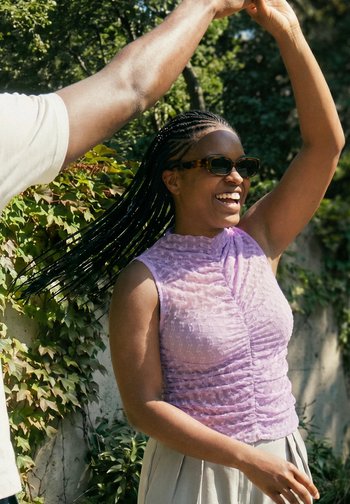 Donna con capelli intrecciati e occhiali da sole sorridente, indossa una canotta viola, balla all'aperto con un braccio sollevato tenuto da un'altra persona.