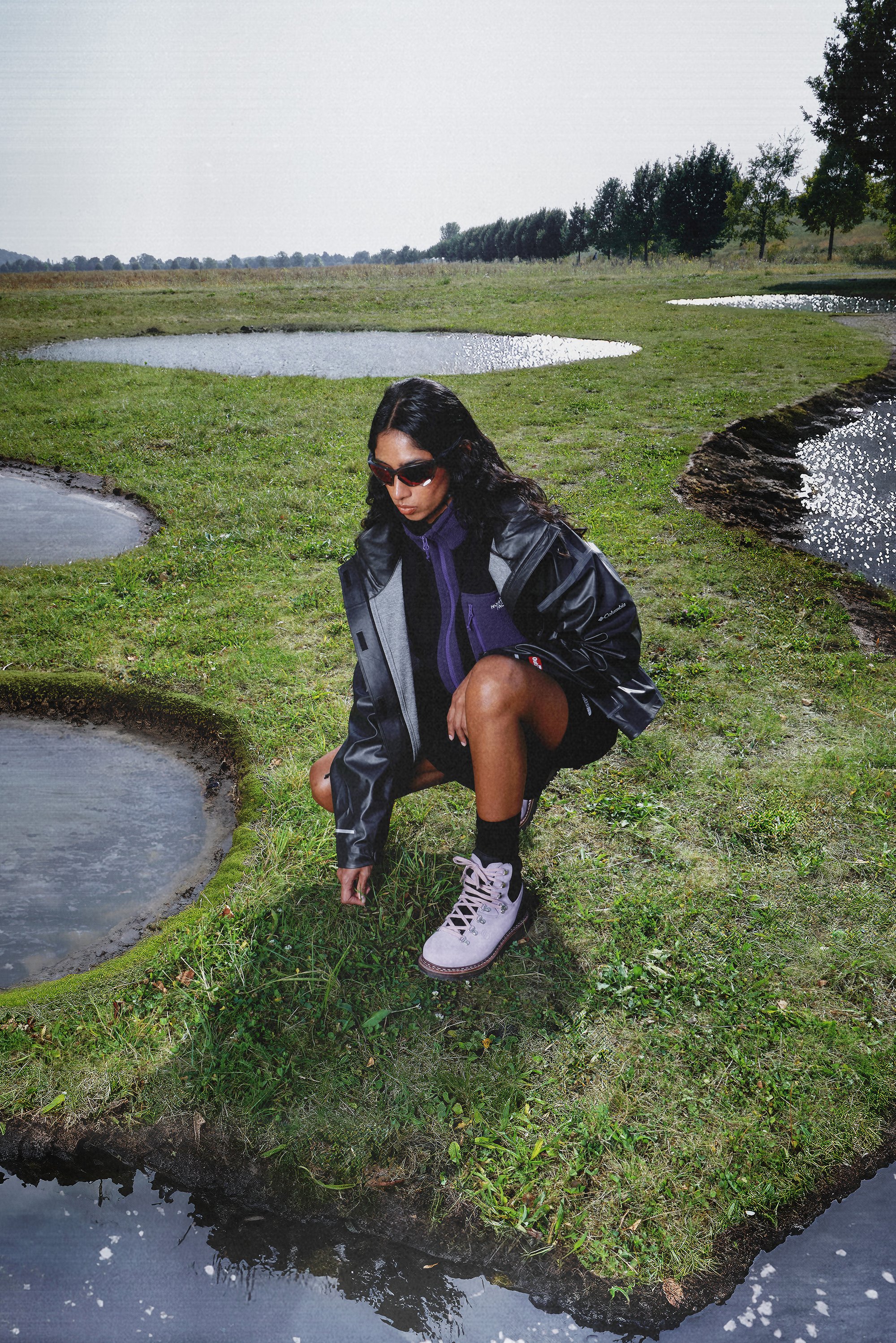 A person in a black jacket and sunglasses crouches by a shallow pond, examining the grass in a grassy field with trees in the background.