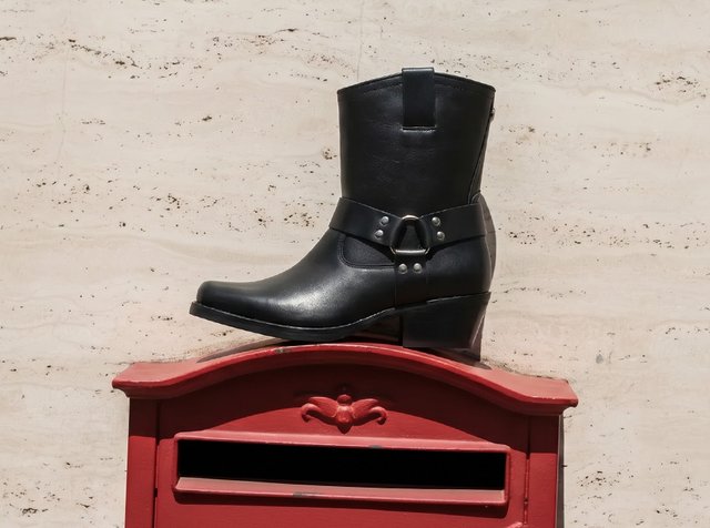 Bottine noire en cuir avec bride décorative et talon, posée sur une boîte aux lettres rouge devant un mur beige.