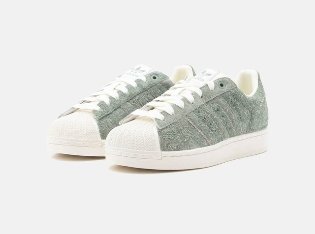 Par de ténis baixos com tecido verde felpudo, biqueiras de borracha branca, atacadores e solas brancas sobre um fundo simples.