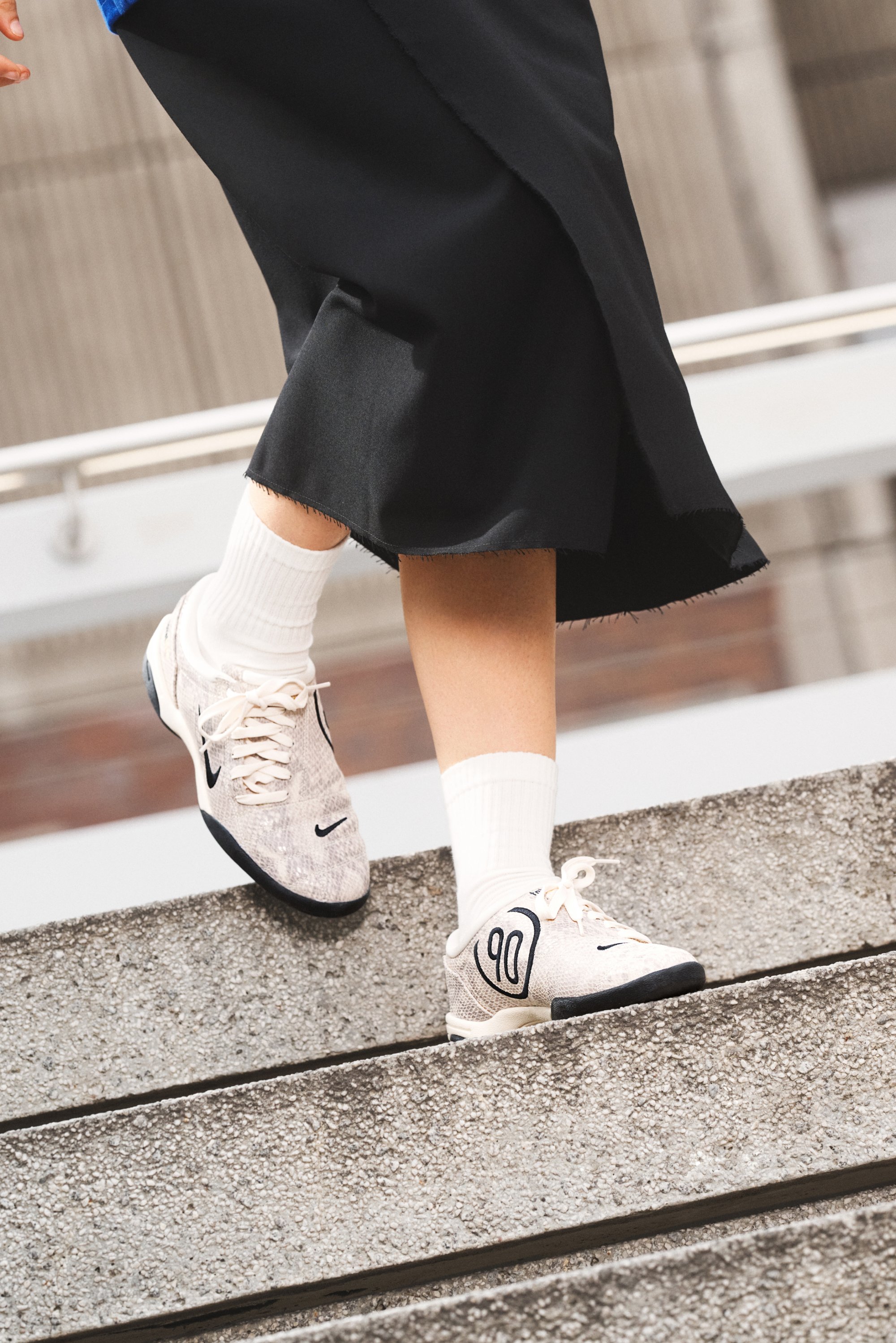 Zapatillas Nike con un upper texturizado ligero y acentos negros, combinadas con calcetines blancos. El modelo desciende por unas escaleras de cemento.