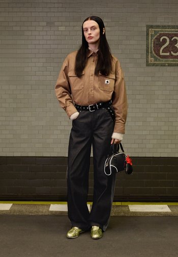Mujer con cabello largo y oscuro de pie frente a una pared de azulejos, vistiendo una camisa marrón, pantalones anchos negros, zapatos verdes y sosteniendo un bolso negro.