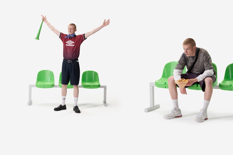 Two young men near green plastic seats; one stands cheering with a green vuvuzela, the other sits eating fries, both wearing casual sportswear.