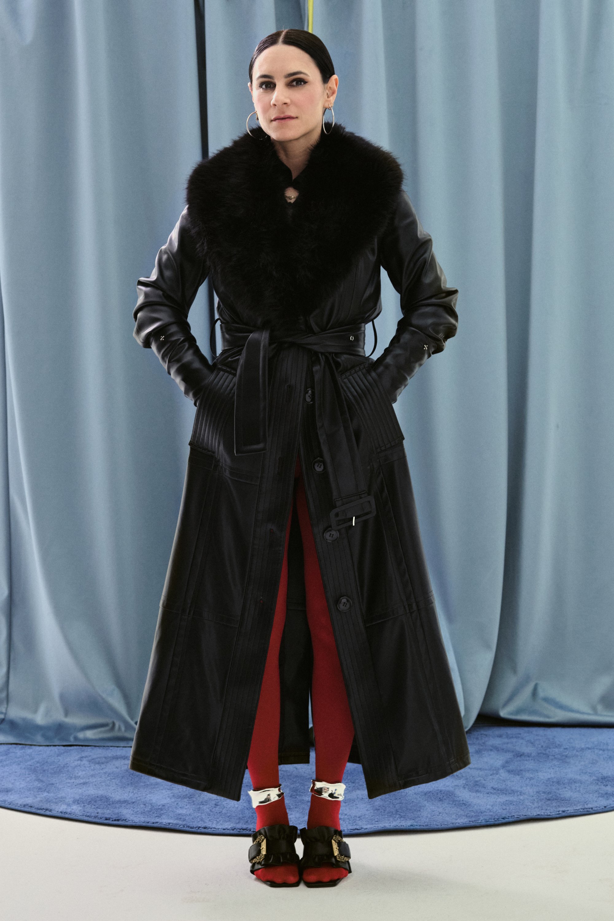Una donna in un lungo cappotto nero con colletto in pelliccia si erge con sicurezza, indossando pantaloni rossi e scarpe uniche, su uno sfondo blu.