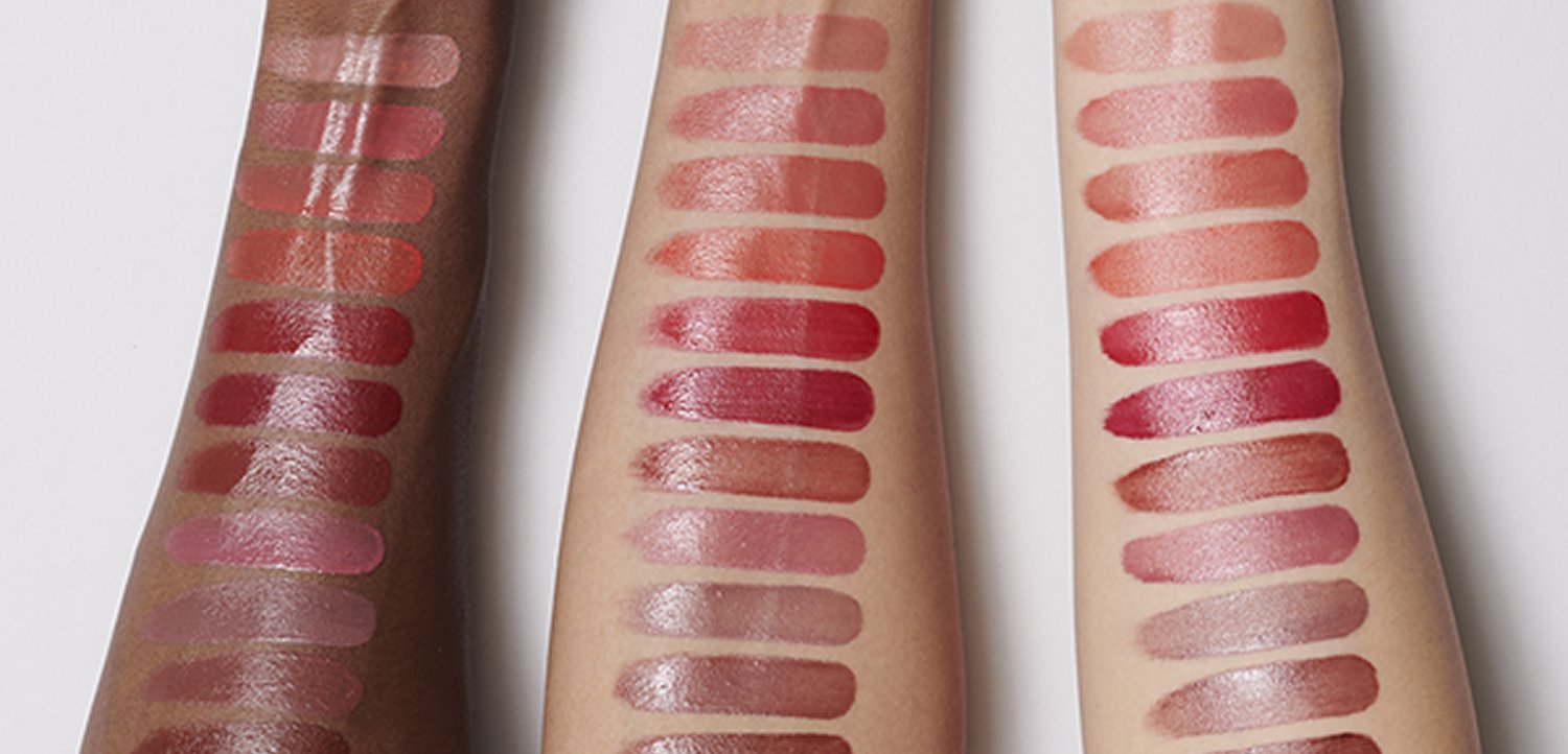 Tre braccia mostrano 12 campioni di rossetto in diverse tonalità di rosso, rosa e nudo; finitura lucida; texture sfumate; tonalità di pelle diverse.