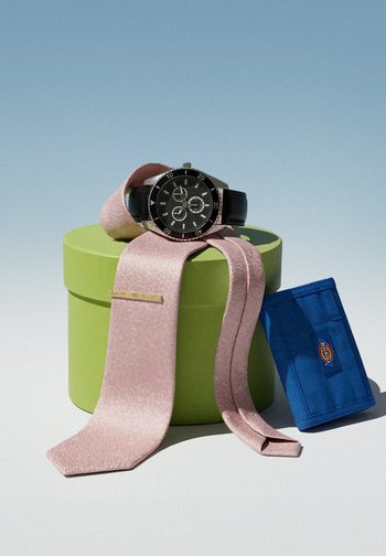 Black wristwatch with leather strap on pink tie, both placed on a green round box, with a blue wallet standing next to the box on a light surface.