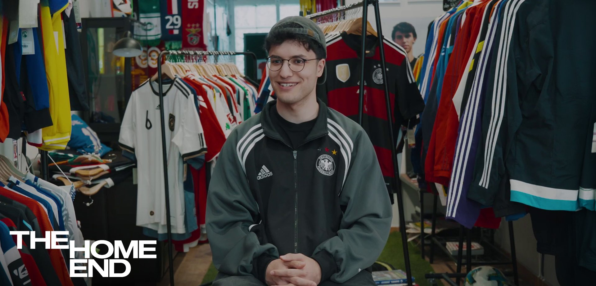 Junger Mann mit Brille und Kappe lächelt, während er in einem Raum sitzt, der mit hängenden Sporttrikots und Kleiderständern gefüllt ist.