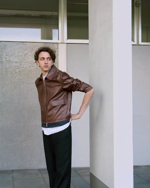 Braune Lederjacke mit Reißverschluss und taillierter Passform, getragen über einem weißen karierten Hemd. Kombiniert mit schwarzen Hosen, die eine lässige Silhouette bieten.