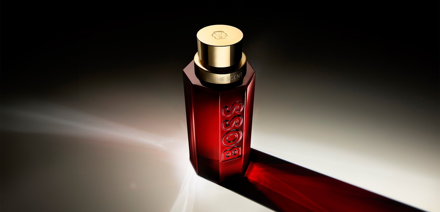 Bottiglia di profumo alta esagonale di colore rosso scuro con tappo dorato e marchio BOSS, che proietta un'ombra lunga su una superficie chiara.