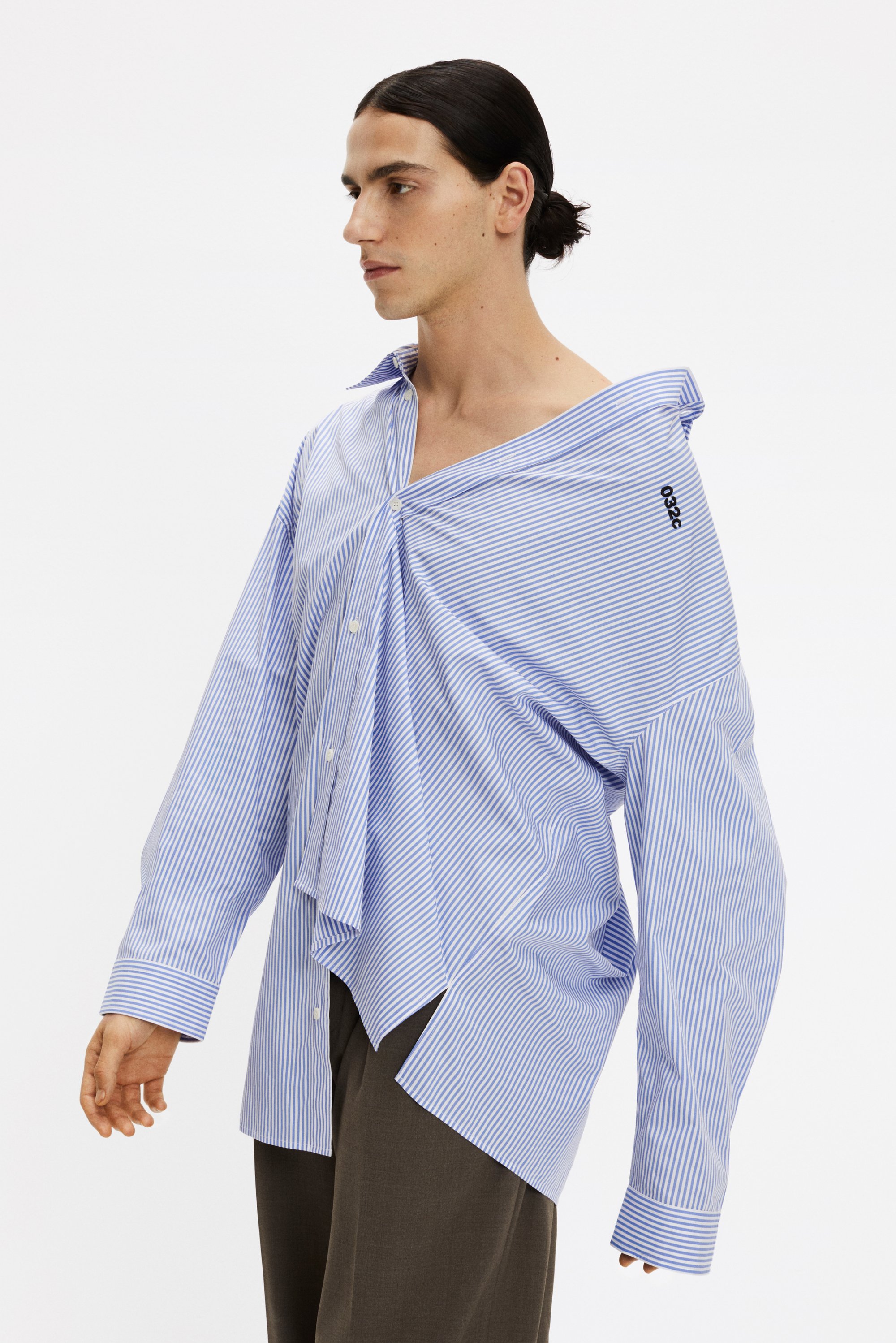 Uomo che indossa una camicia a righe blu e bianche, oversize e sbottonata, abbinata a pantaloni scuri, in piedi contro uno sfondo bianco semplice.