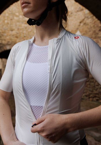 Una donna sistema la cerniera della sua maglietta da ciclismo bianca, in piedi in un ambiente rustico con muri di pietra e luce naturale.