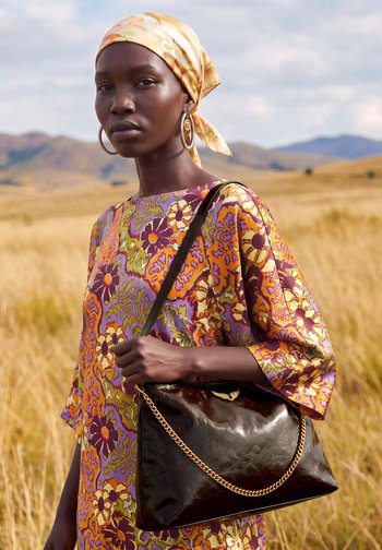 Femme portant de grandes boucles d'oreilles en anneau et un foulard jaune sur la tête, debout dans un champ, vêtue d'une robe fleurie colorée et tenant un sac à main en cuir sombre.