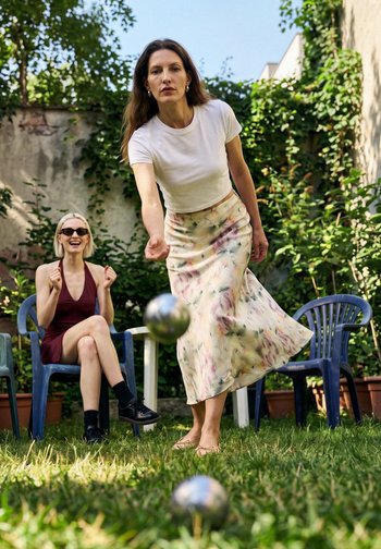 Donna con camicia bianca e gonna floreale lancia una palla di metallo mentre un'altra donna con occhiali da sole osserva seduta su una sedia blu all'aperto.