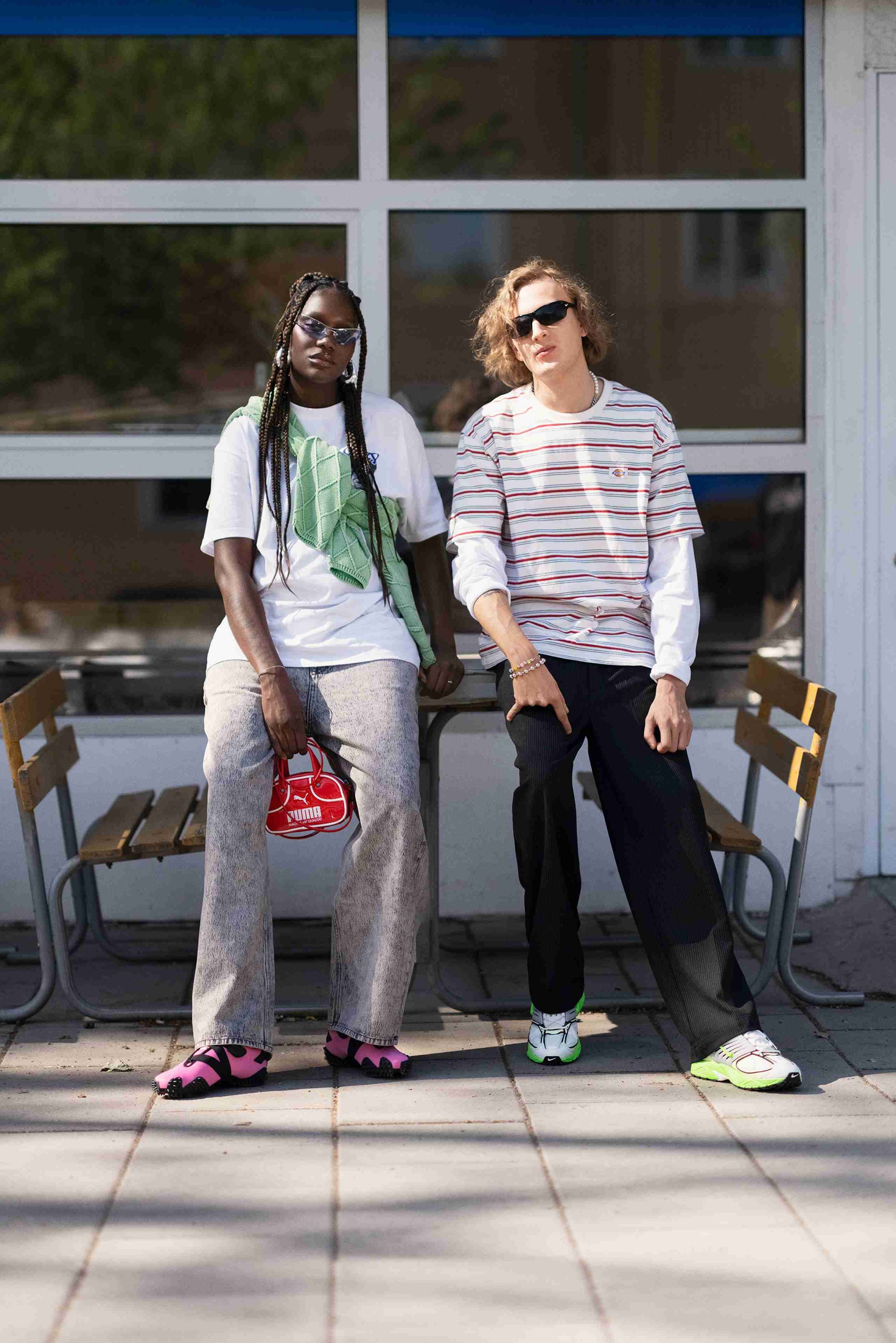 Twee personen zitten op een bank. De ene draagt een wit T-shirt, een groene trui, lichte spijkerbroek en roze schoenen. De ander heeft een gestreept shirt, zwarte broeken en groene accenten op de schoenen.