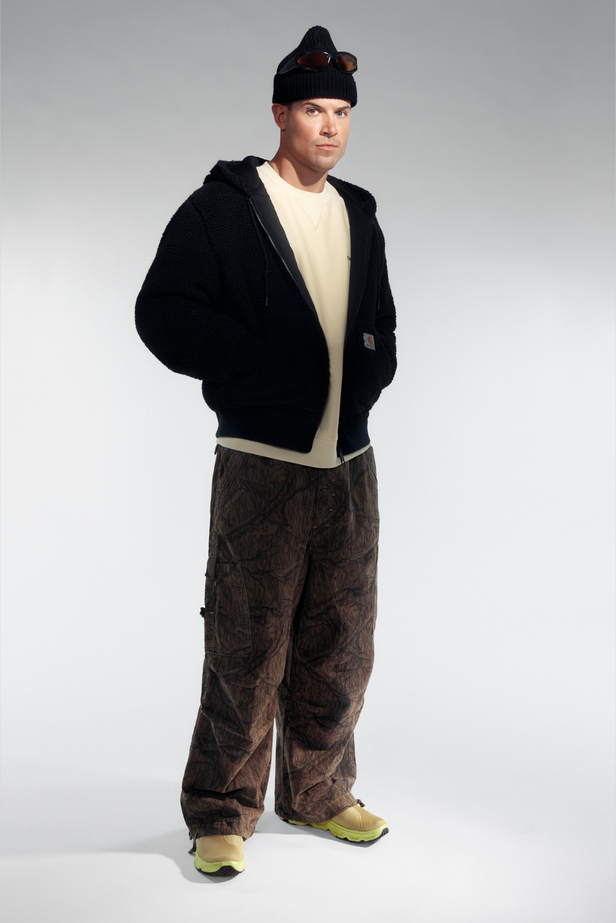 Un hombre se presenta con confianza en una cómoda sudadera negra, camisa beige, pantalones marrones texturizados y zapatillas amarillas brillantes, con un gorro y gafas de sol.