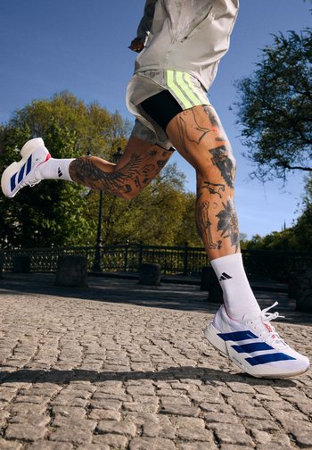 Tatoveret løber i hvide Adidas-sneakers og stribede shorts midt i et skridt på en solrig brostenssti, med træer og parkhegn i baggrunden.