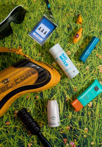 Un sac de sport jaune, des lunettes de soleil, une brosse à cheveux et divers produits de soin de la peau sont disposés sur une surface à motif herbe vert.