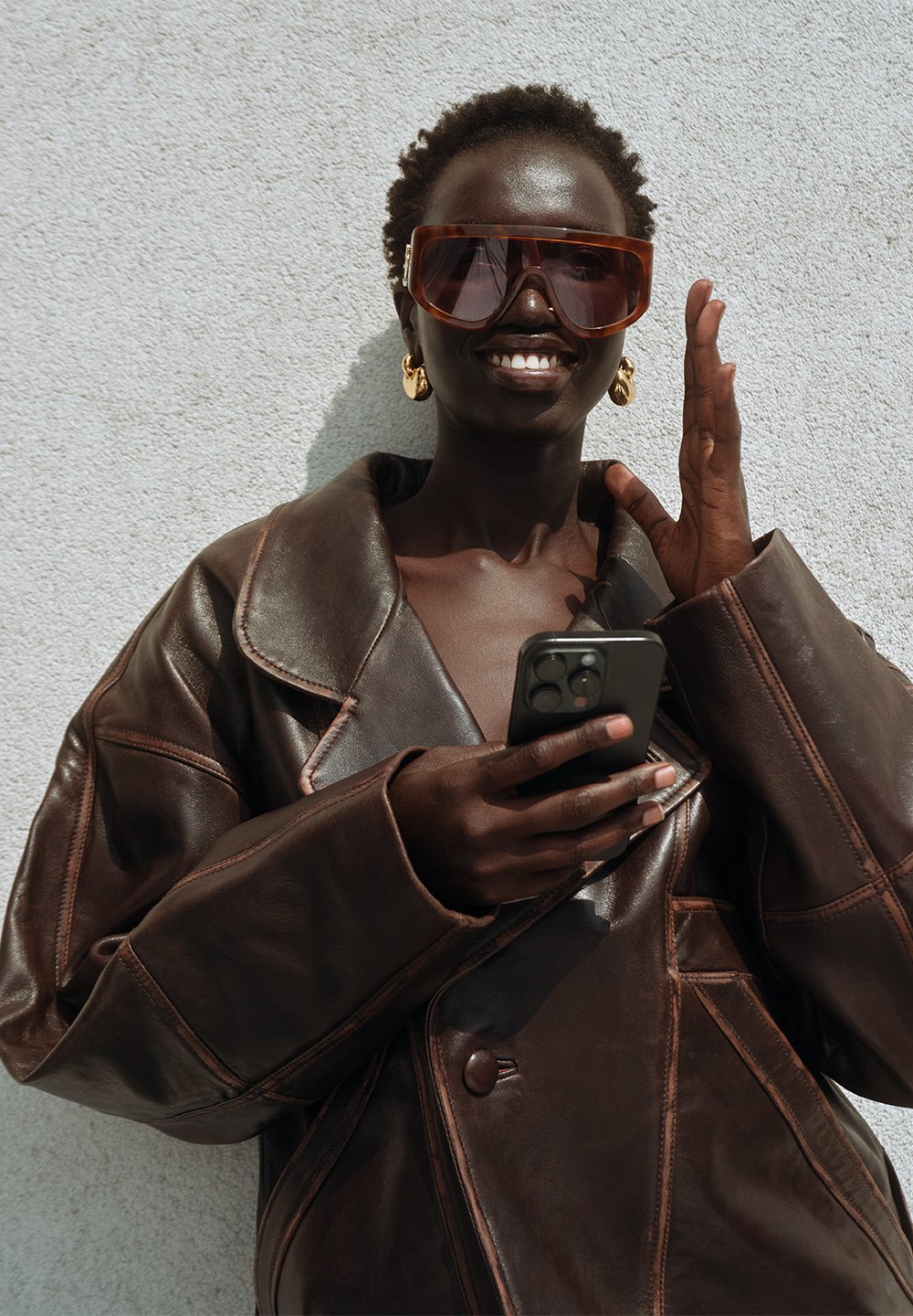 Brown leather oversized jacket featuring a structured collar, contrasting stitching, and prominent buttons. Complemented by large orange-tinted sunglasses.