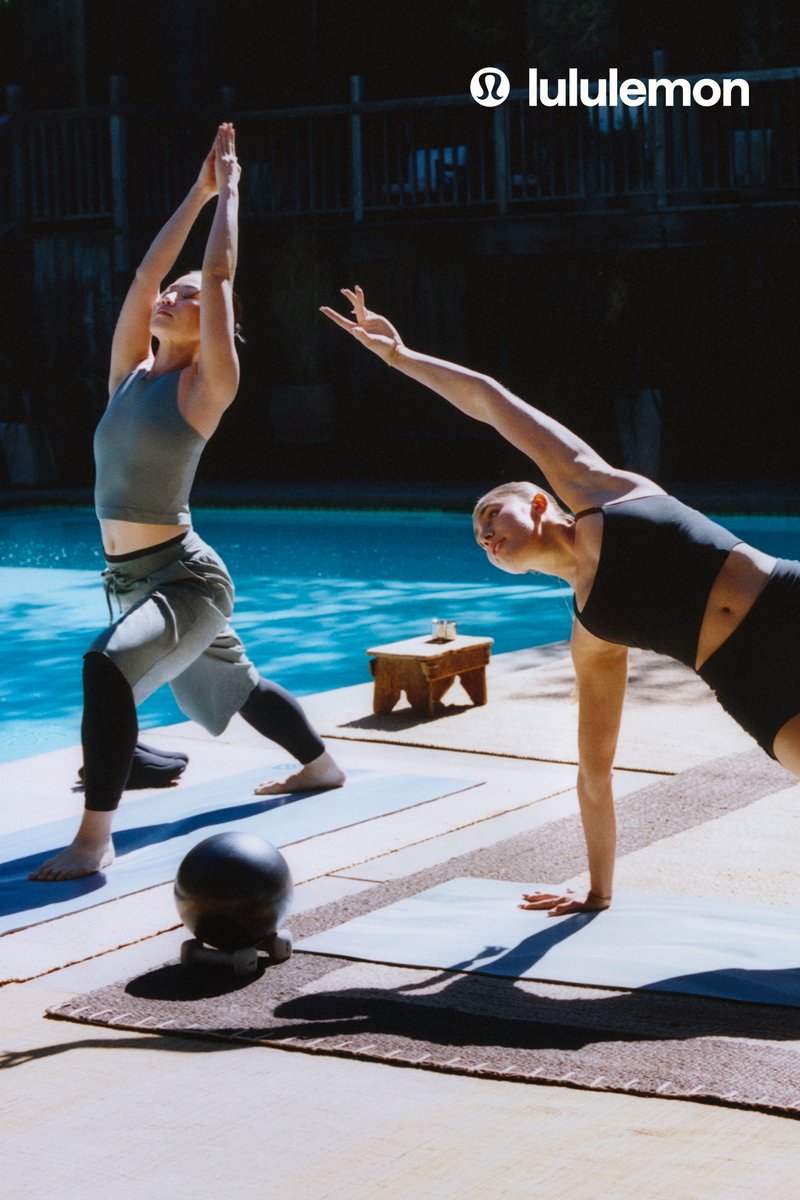 Two women practice yoga by a pool, one in a side plank and the other in a raised arm stretch, both on mats with yoga gear nearby.