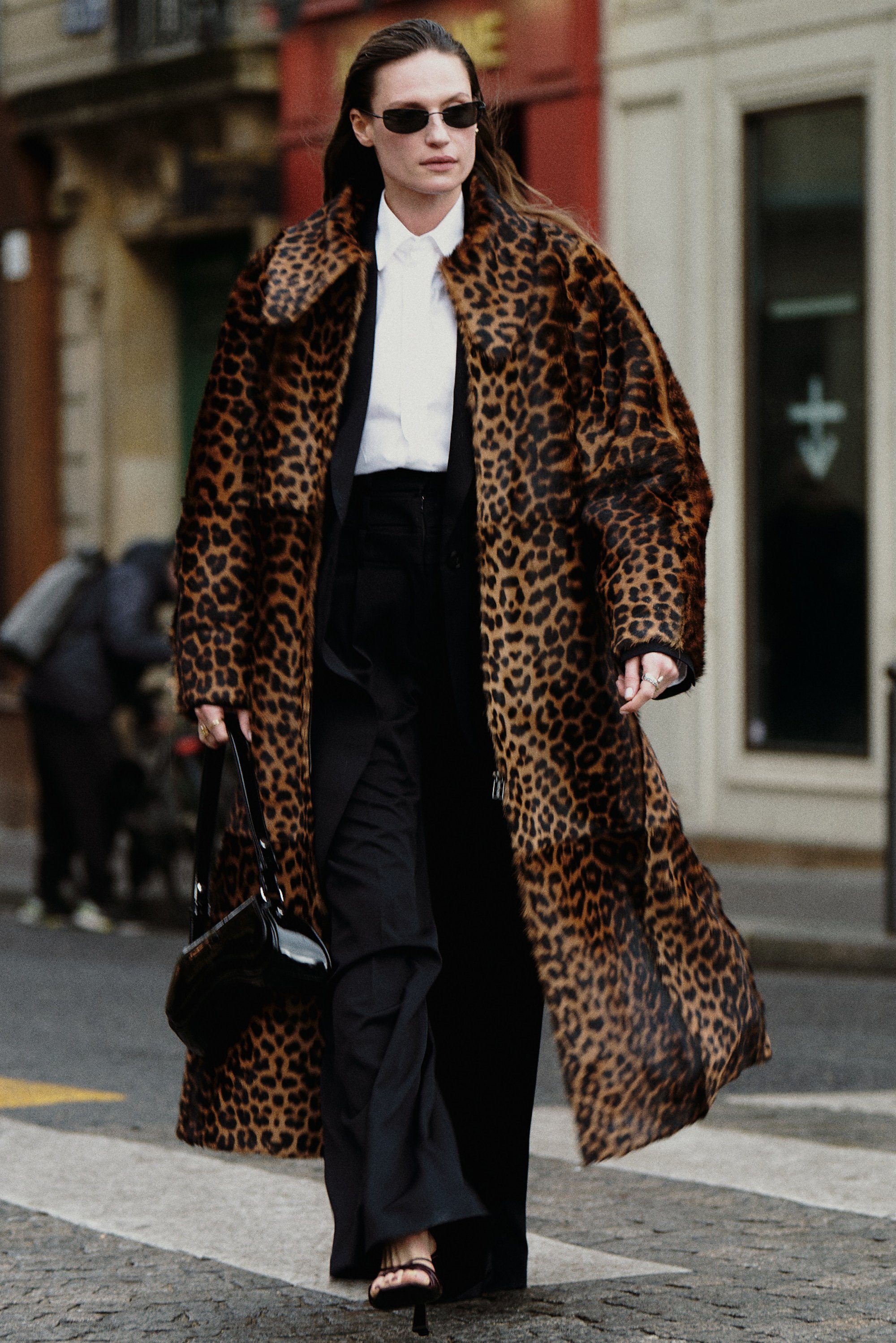 Una mujer camina con confianza por una calle de la ciudad, luciendo un abrigo de leopardo, pantalones negros, gafas de sol y llevando un bolso pequeño.
