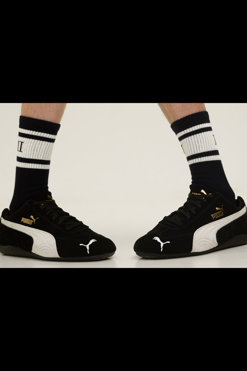 Schwarze Wildledersneaker mit weißen Akzenten und goldenen Logo; kombiniert mit schwarz-weißen gestreiften Socken. Die Sohle verfügt über strukturierte Grip-Muster.
