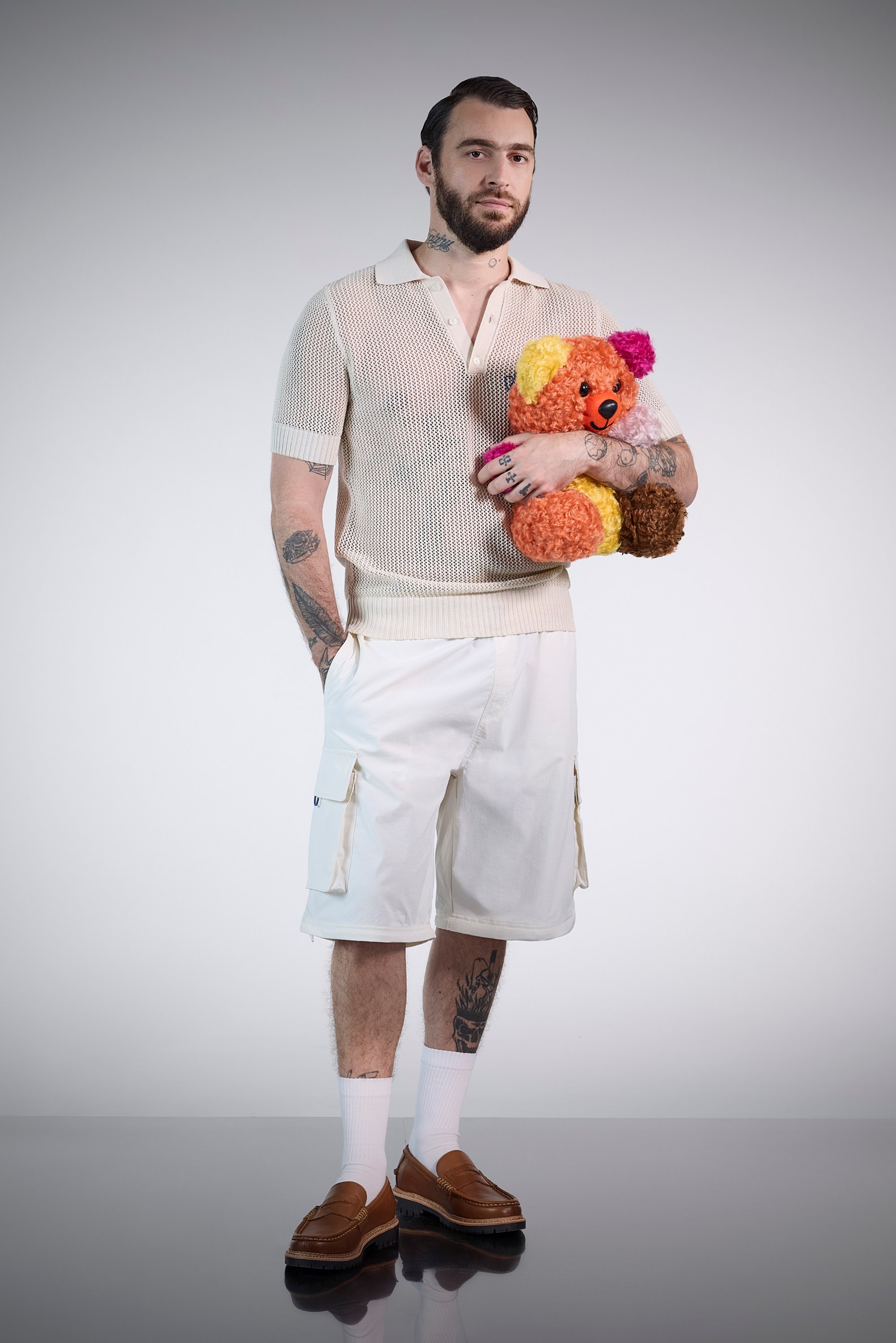 Man with a beard and tattoos wears a knitted polo shirt and cargo shorts, holding a colourful teddy bear, set against a plain background.