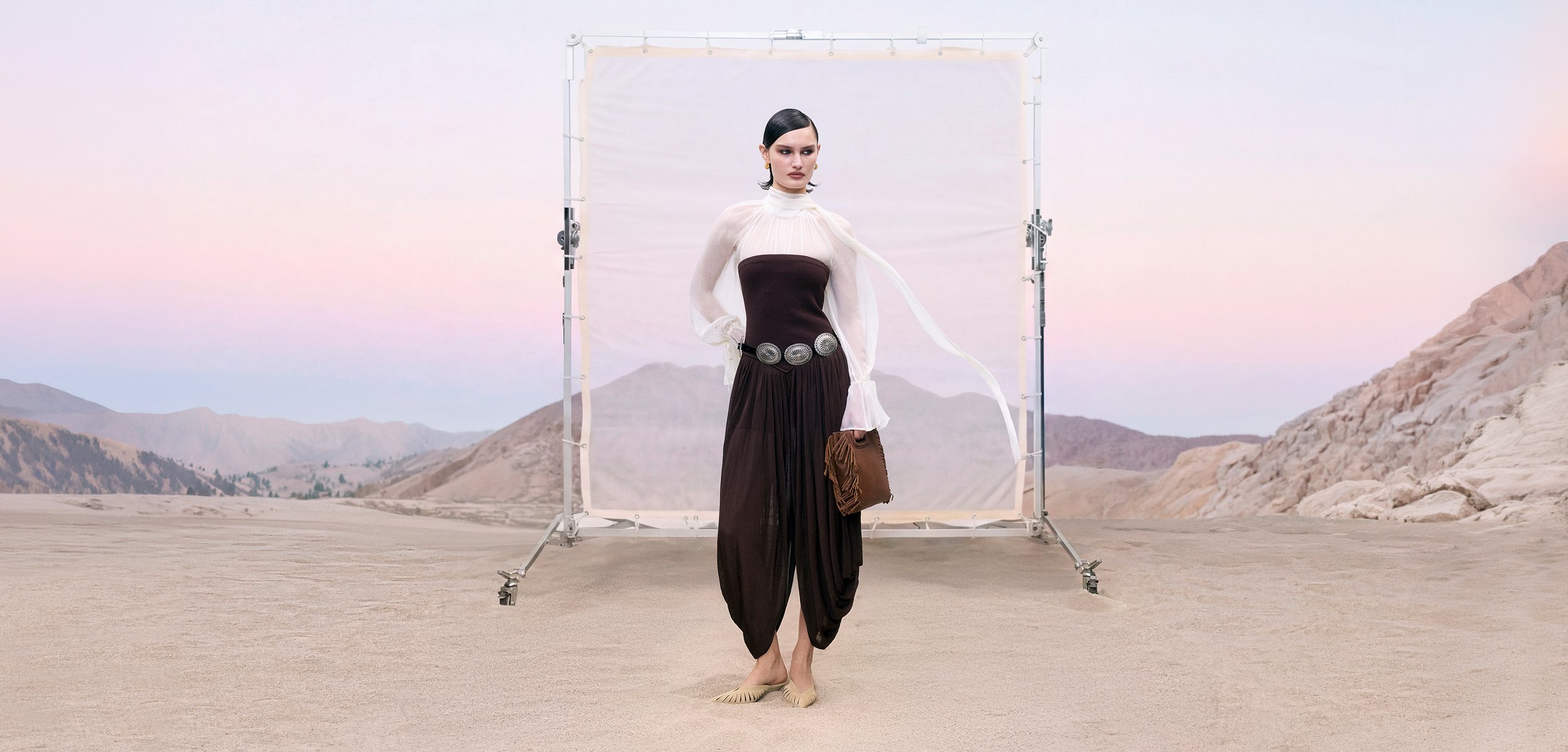 Donna in outfit di alta moda con top bianco trasparente e tuta nera senza spalline, che tiene una pochette marrone, in piedi a piedi nudi sulla sabbia del deserto con montagne sullo sfondo.
