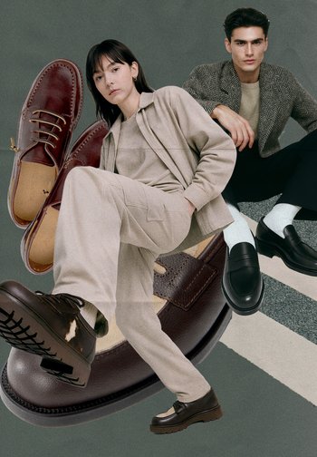 Bruna läderskor med snören och en slät svart loafers på en strukturerad yta, bredvid personer i kläder i neutrala toner.