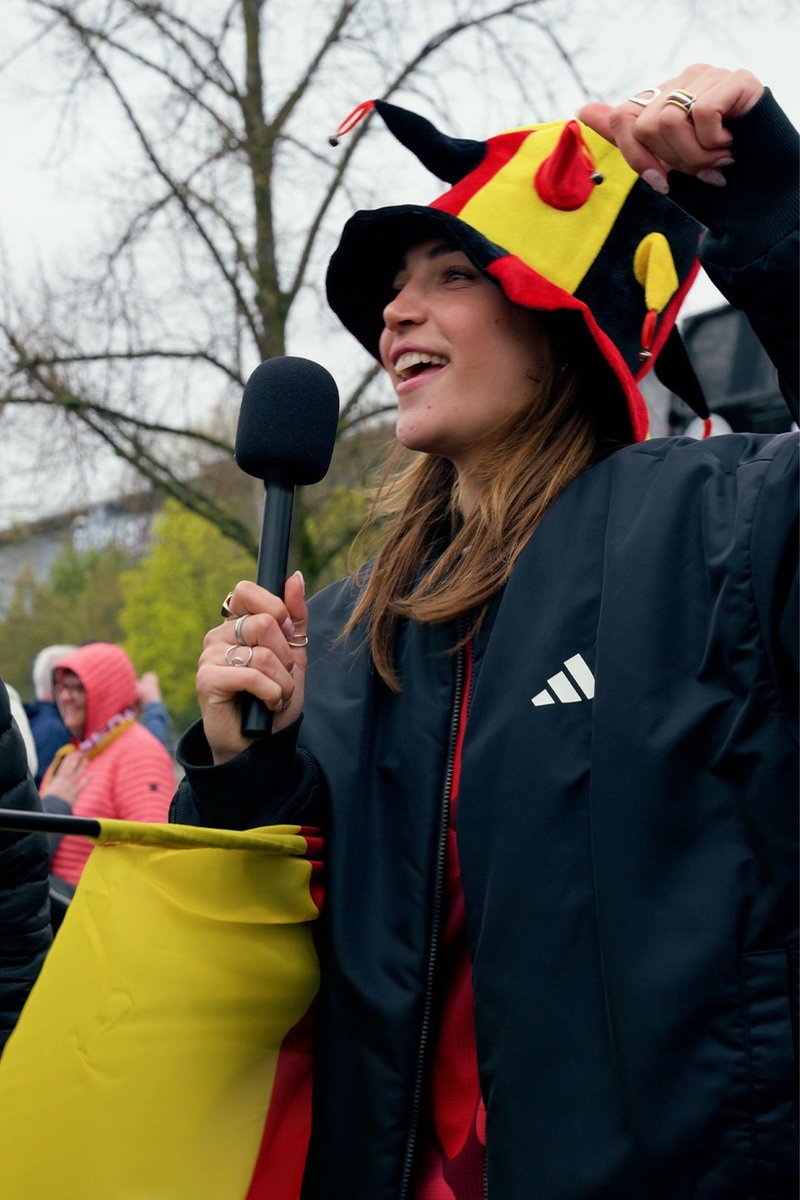 Woman wearing a red, yellow, and black jester hat holds a microphone and matching flag while speaking outdoors at an event.