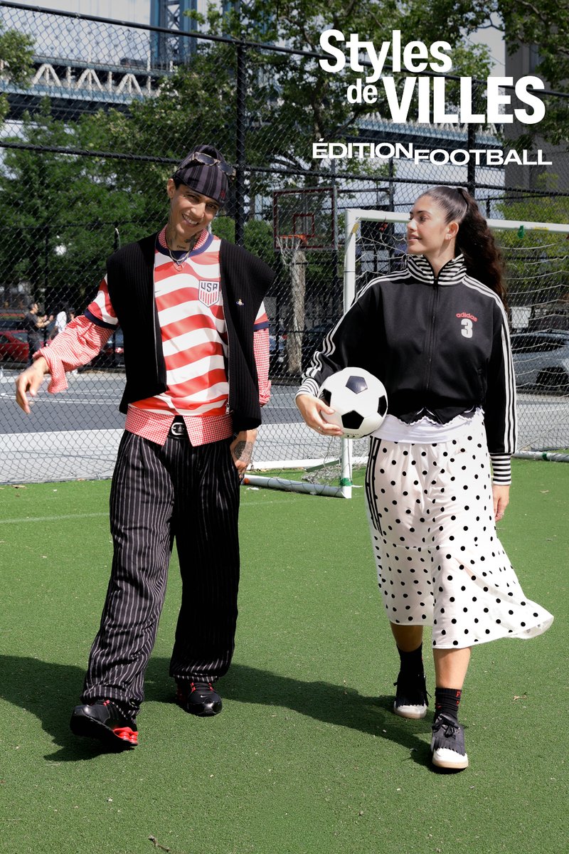 Deux personnes marchent sur un terrain de football, l'une tenant un ballon de football, avec un but et une clôture en arrière-plan sous des arbres et un pont.