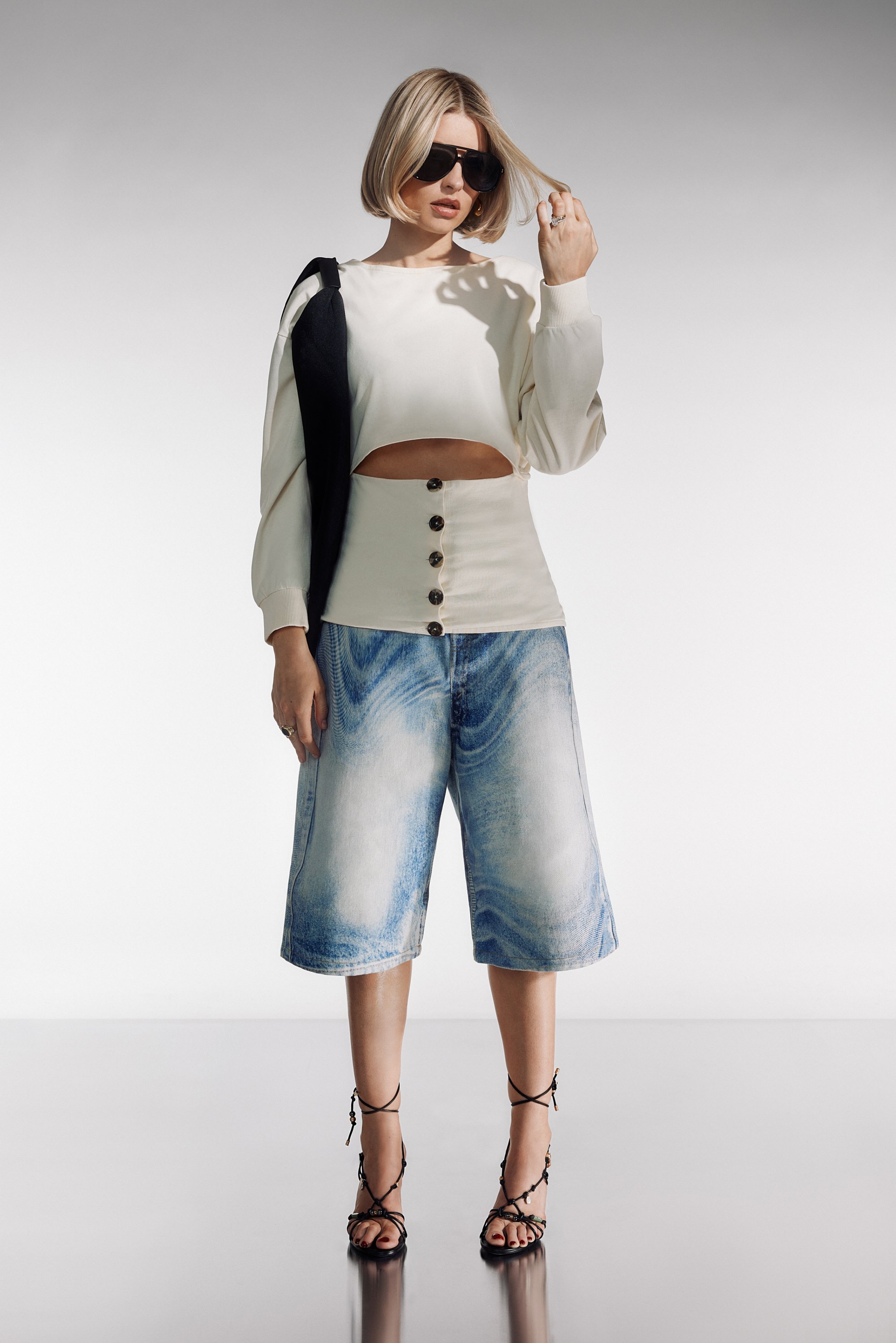 Una mujer con cabello rubio corto lleva una blusa corta color crema, pantalones cortos de mezclilla de tiro alto y tacones negros con tiras, adoptando una pose segura.