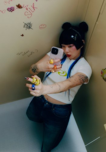 Una persona joven con cabello azul y tatuajes sostiene un teléfono, posando con confianza. El fondo presenta graffiti y garabatos en una pared.