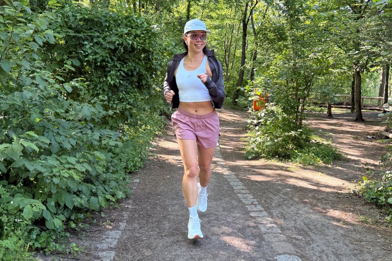Femme courant sur un sentier forestier ombragé, portant une casquette blanche, un short rose, des baskets blanches et des lunettes de soleil, souriant par temps ensoleillé.