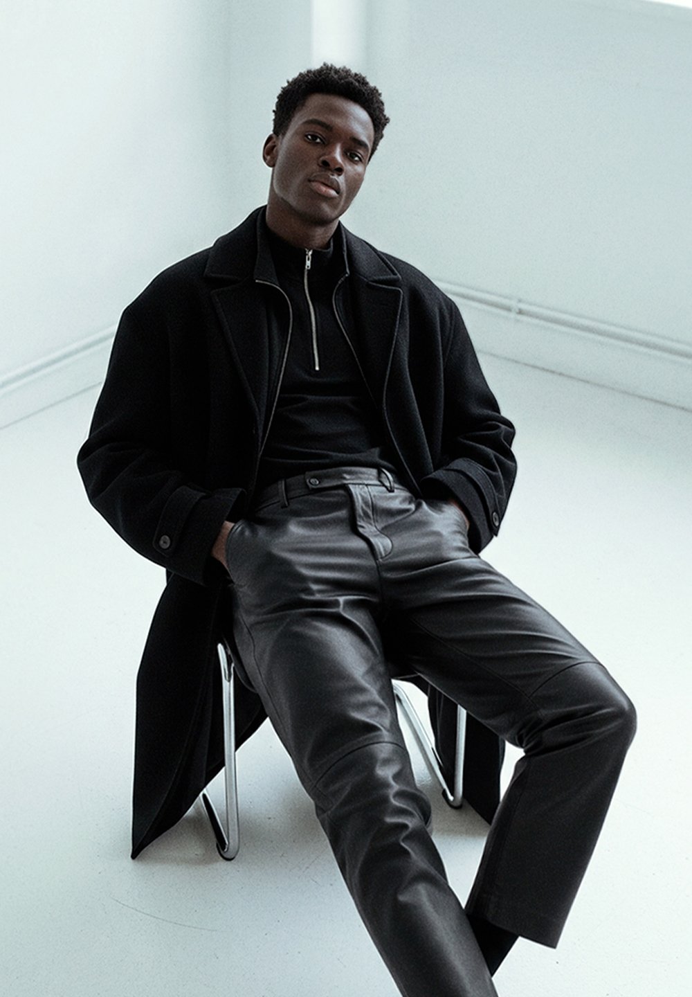 Black wool overcoat layered over a black zip-up top and leather trousers, seated on a metallic chair against a light background.