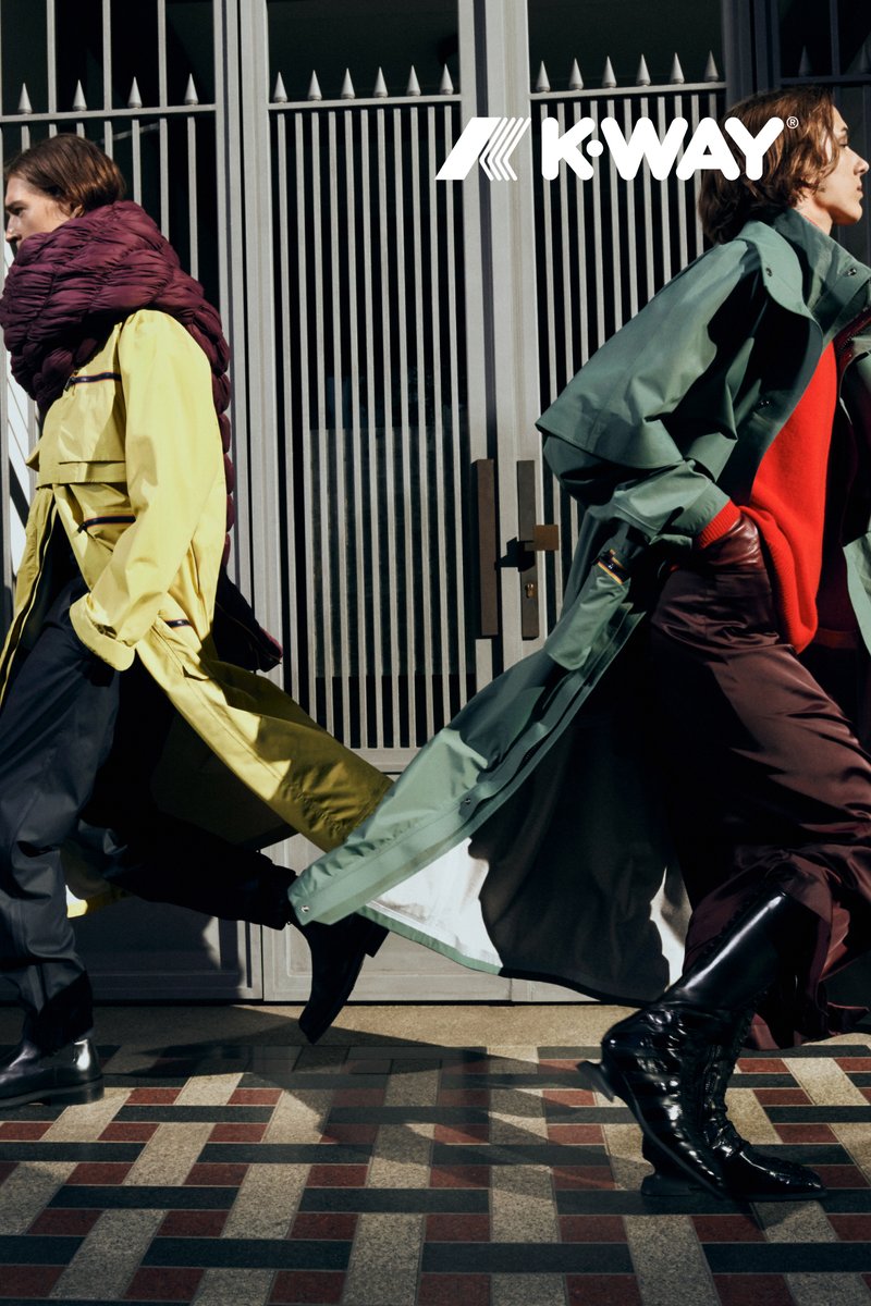 Zwei Models laufen in einer Stadtkulisse, bekleidet mit einem gelben Trenchcoat und einem grünen Mantel, ergänzt durch einen auffälligen roten Pullover und dunkle Hosen.